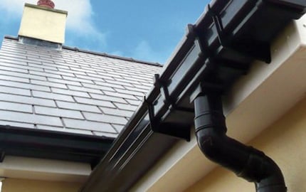 Black gutter and downspout system installed on a house roof with chimney visible against blue sky