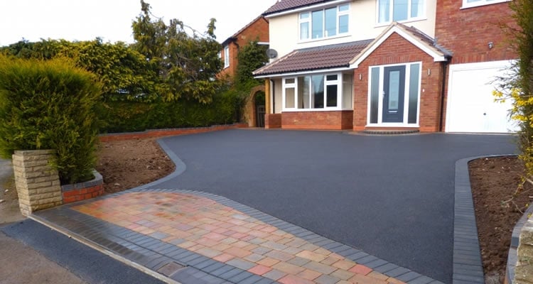 Modern brick house with dark driveway and paved entrance, bordered by landscaping and shrubs