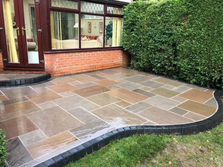 Curved patio with mixed stone tiles, bordered by dark edging, adjacent to brick house with ivy-covered wall and green lawn