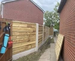 Wooden fence panels along a narrow pathway between red brick buildings and residential garden area