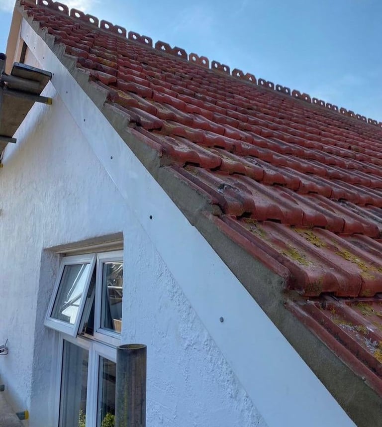 White-walled building with arched window beneath a steep roof covered in red clay tiles showing moss growth, against a clear blue sky
