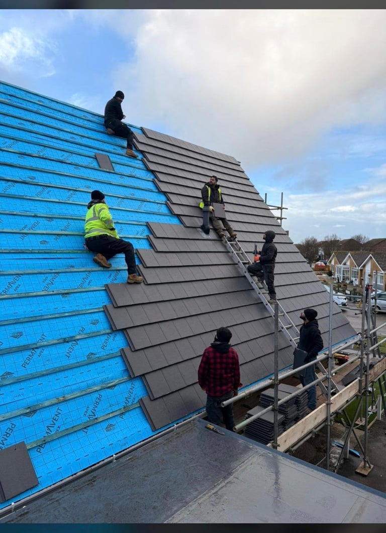 Construction workers installing gray roof tiles on a residential building, with blue protective sheeting visible on the left side