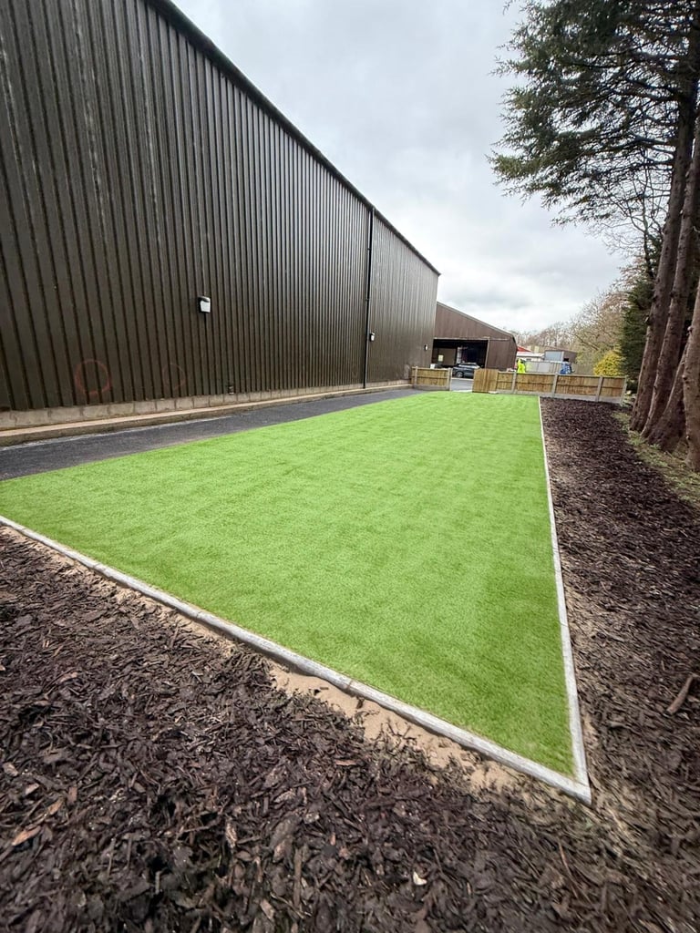 Bright green artificial turf courtyard bordered by mulch beside a large corrugated metal building with trees in background